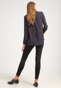 Donna con capelli lunghi che indossa una blusa a maniche lunghe a motivo navy, pantaloni neri aderenti e scarpe nere con tacco basso, in piedi su un pavimento bianco.