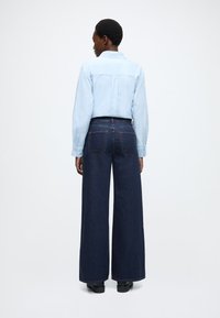 Svetlomodrá košeľa na gombíky s dlhými rukávmi, doplnená o tmavé džínsy s vysokým pásom a širokými nohavicami. Viditeľná je čierna obuv.