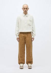 Ανοιχτόχρωμο πουκάμισο με ρίγες και μπροστινή τσέπη, συνδυασμένο με φαρδιά καφέ παντελόνια από κορντούρα. Το μοντέλο στέκεται σε ουδέτερο φόντο.
