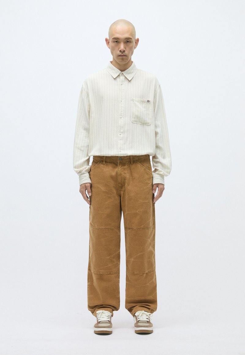 Ανοιχτόχρωμο πουκάμισο με ρίγες και μπροστινή τσέπη, συνδυασμένο με φαρδιά καφέ παντελόνια από κορντούρα. Το μοντέλο στέκεται σε ουδέτερο φόντο.
