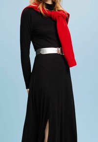 Schwarzes langärmeliges Kleid mit einem hohen Schlitz, kombiniert mit einem roten gestrickten Schal und einem silbernen metallischen Gürtel, vor einem hellblauen Hintergrund.