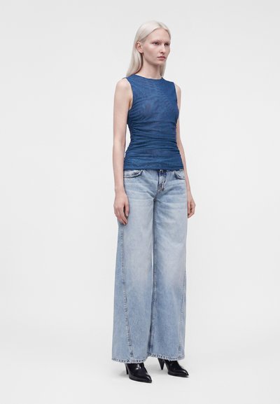 Femme aux longs cheveux blonds portant un haut bleu sans manches, un jean large bleu clair et des talons noirs pointus, debout devant un fond uni.