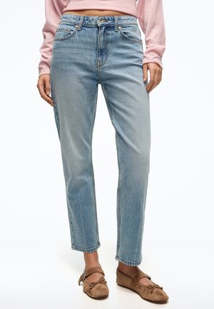 Frau trägt hellblaue Jeans mit geradem Bein, ein langärmliges pinkes Oberteil und braune Wildleder-Mary-Jane-Schuhe mit Schleifen.