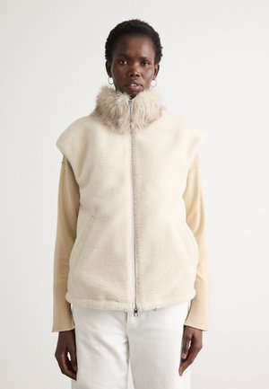 Een vrouw draagt een crèmekleurige mouwloze vachtvest met een hoge bontkraag over een beige langemouwtop en witte broek, staand tegen een witte achtergrond.