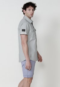 Camicia grigia chiaro a maniche corte con due tasche sul petto, colletto button-down e toppa con logo sulla manica sinistra; abbinata a pantaloni corti leggeri di colore viola chiaro arrotolati.