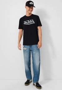 Muž se usmívá, nosí černou kšiltovku Von Dutch a tričko, světle modré džíny s nášivkami a černé tenisky s bílými pruhy.