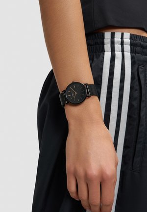 Zwarte horlogeband van mesh met een minimalistische ronde wijzerplaat, voorzien van gouden uurmarkeringen en wijzers, gedragen om een pols tegen een achtergrond van zwarte sportshorts.