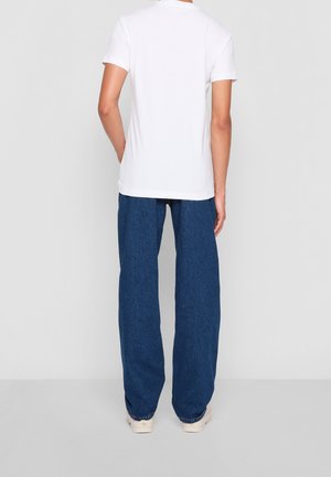 Person trägt ein schlichtes weißes T-Shirt mit kurzen Ärmeln, lockere blaue Jeans und weiße Sneakers, steht mit dem Rücken zu einem schlichten Hintergrund.