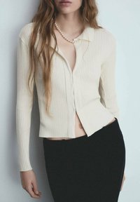 Cardigan a coste di colore crema con maniche lunghe, scollatura a V e bottoni; indossato sopra una gonna nera. Presenta un design corto e cuciture a vista.