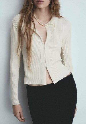 Cardigan a coste di colore crema con maniche lunghe, scollatura a V e bottoni; indossato sopra una gonna nera. Presenta un design corto e cuciture a vista.