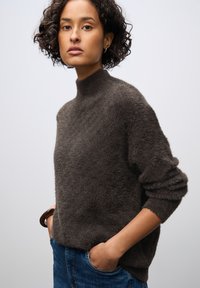 Maglione grigio scuro, peloso, con collo alto e vestibilità oversize; abbinato a jeans in denim blu; esposto su uno sfondo neutro.