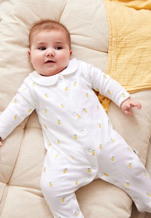 Bebé acostado sobre un cojín color crema que lleva un body blanco con pequeñas piñas amarillas y un cuello con borde festoneado.