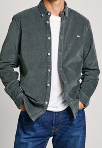 Camicia in velluto a coste grigia con colletto a bottoni, tasche frontali e dettaglio del logo bianco. Indossata sopra una T-shirt bianca, abbinata a jeans blu.