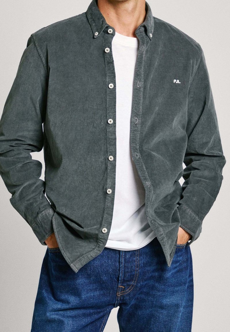 Camicia in velluto a coste grigia con colletto a bottoni, tasche frontali e dettaglio del logo bianco. Indossata sopra una T-shirt bianca, abbinata a jeans blu.