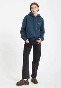 Teal hoodie with drawstrings, front logo, paired with black wide-leg jeans and brown lace-up shoes. Background is white.