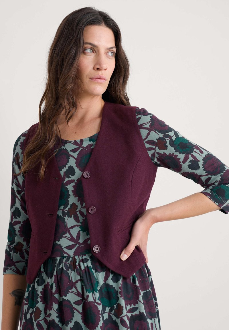 Burgundski vuneni prsluk s krojem, s pet dugmadi i bez ovratnika, nosi se preko haljine s uzorkom u zeleno-burgundskim cvjetnim motivima.