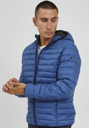 Chaqueta acolchada azul con capucha, que presenta un diseño acolchado y un zipper delantero. El material parece suave y ligero.