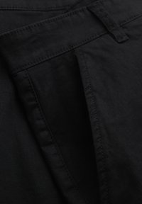 Gros plan sur un vêtement en tissu noir montrant une poche et une boucle de ceinture avec des coutures détaillées.