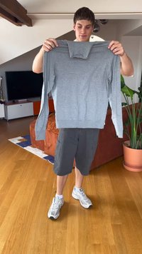 Młody mężczyzna trzyma szary sweter z długim rękawem w pomieszczeniu, stojąc na drewnianej podłodze obok pomarańczowej kanapy i doniczki z rośliną.