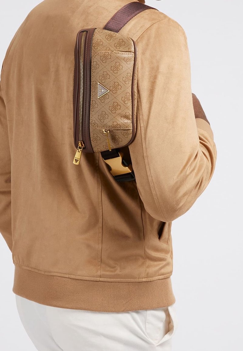 Homme portant une veste en daim couleur beige et un pantalon blanc, avec un sac bandoulière marron à motifs fermé par une fermeture éclair dans le dos.
