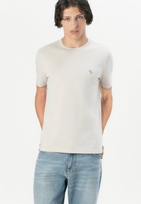 T-shirt en coton gris clair à manches courtes, col rond, avec un petit logo brodé sur la poitrine. Associé à un jean en denim bleu.