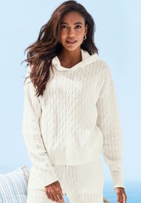 Gestrickter Kapuzenpullover in Weiß mit einem strukturierten Zopfmuster. Mit gerippten Bündchen und Saum ausgestattet. Kombiniert mit passenden Hosen. Heller Hintergrund.