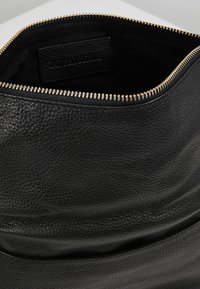 Bolso de mano de cuero negro con un acabado texturizado. Presenta un zipper en tono dorado y una etiqueta interior. El diseño es minimalista y funcional.