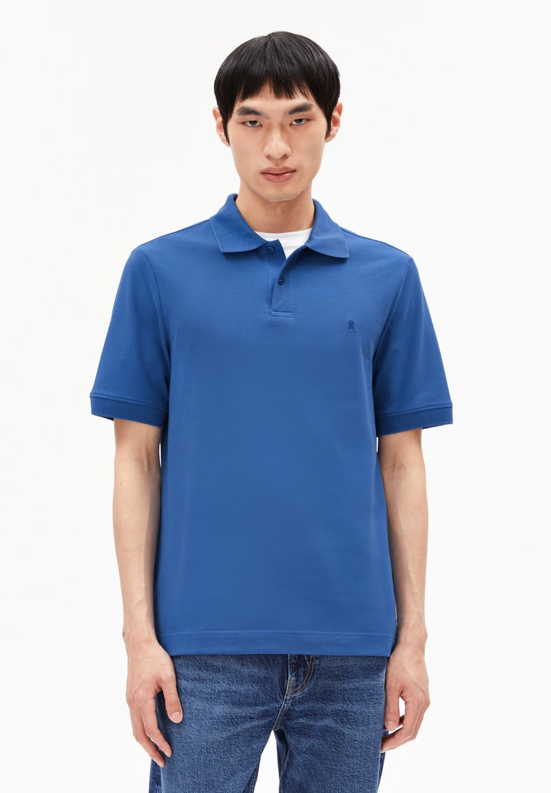 Blaues Poloshirt aus Baumwoll-Mischung, mit classicem Kragen, kurzen Ärmeln und kleinem Logo auf der Brust. Getragen mit Denim-Jeans.