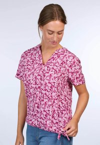 Kurzarmpullover mit V-Ausschnitt und einem pinken Blumenmuster auf leichtem Stoff. Mit einem gebundenen Detail an der Taille. Lässige Passform, geeignet für warmes Wetter.