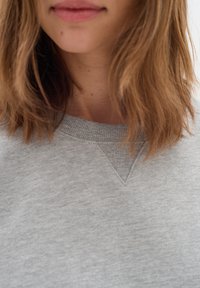 Mujer con cabello castaño claro hasta los hombros que lleva un suéter gris claro de cuello redondo, mostrando solo la parte inferior del rostro y el cuello.