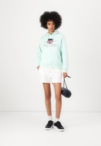 Mintgroene hoodie met logo, kangoeroezak, en trekkoord. Gecombineerd met witte shorts en zwarte sneakers; vasthoudend een kleine zwarte tas.