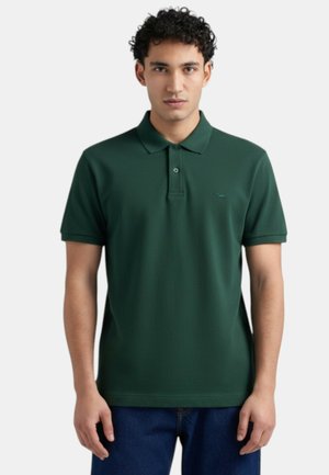 Giovane uomo che indossa una polo verde scuro a maniche corte e jeans blu, in piedi contro uno sfondo chiaro e semplice.