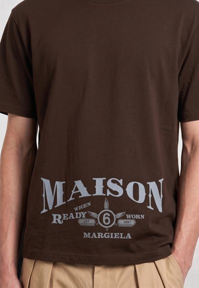 Καφέ μπλουζάκι με μεγάλο λευκό κείμενο "MAISON READY WHEN WORN" και "6 MARGIELA" τυπωμένα κοντά στο κάτω εμπρός μέρος, φορεμένο από ένα άτομο.