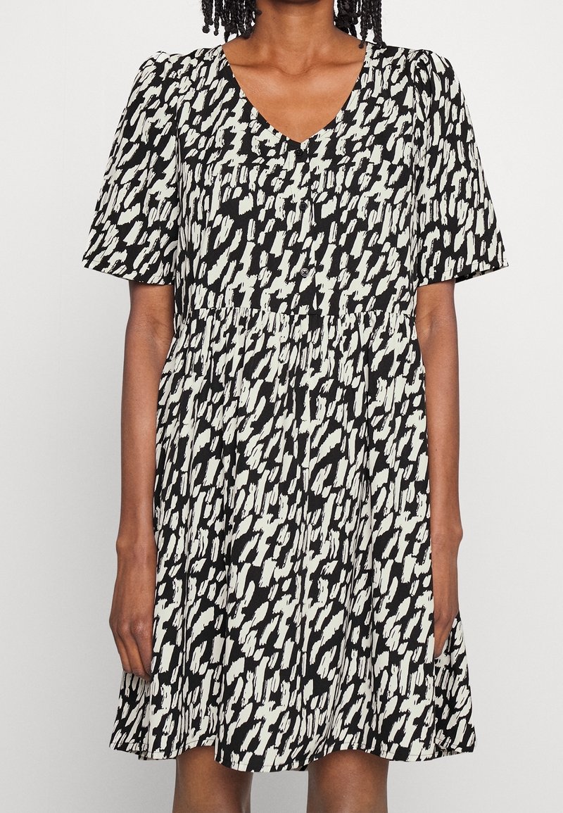 Femme portant une robe à manches courtes avec un motif noir et blanc, un col en V et des boutons sur le devant.
