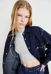 Mørk denimjakke med hvid syning, knaplukning og sidelomme, båret over en lys grå crop top og mørke jeans.
