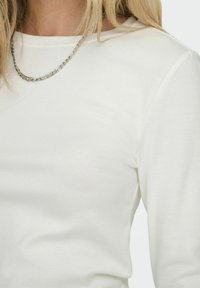Haut blanc à manches longues en tissu tricoté doux avec un col rond, arborant une texture subtile. Accompagné d'un collier en chaîne argentée.
