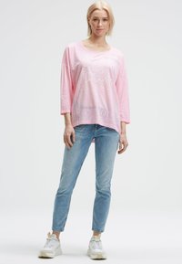 Woman with short blonde hair wearing a loose pink long-sleeve top with text, light blue jeans, and white sneakers, standing against white background.
