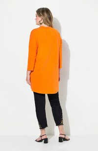 Túnica de manga larga naranja con un corte relajado, combinada con jeans oscuros con detalles dorados en el dobladillo y sandalias negras de tacón.