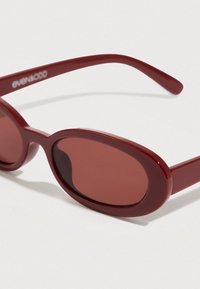 Κόκκινα οβάλ γυαλιά ηλίου με σκούρους φακούς. Κατασκευασμένα από γυαλιστερό πλαστικό με λεπτά μπράτσα, φιλοξενούν το όνομα της μάρκας "evenodd" στον βραχίονα.