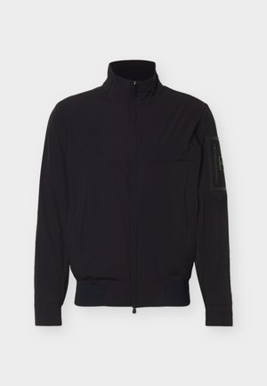 Veste noire à fermeture éclair avec col montant, manches longues, taille élastique et une écusson rectangulaire sur le bras gauche supérieur.