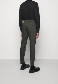 Man med kolgrå slim-fit byxor, svart skjorta, svarta strumpor och svarta läder monk strap-skor, som står framför en enfärgad bakgrund.