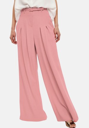 Donna che indossa pantaloni rosa a vita alta, a gamba larga, con pieghe e cintura abbinata, abbinati a un top bianco e tacchi aperti color bronzo.