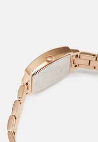 Rose gold metal ur med et rektangulært urkasse, en poleret finish og en tekstureret krone. Har en led-armbånd med en glat tekstur.