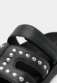 Μαύρο δερμάτινο σανδάλι με δύο πλατιές λουρίδες, διακοσμητικά με τρουκ και σχέδιο ανοιχτού δαχτύλου. Μέγεθος σημειωμένο ως 37. Λεία υφή με αντίθετες λεπτομέρειες.