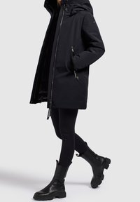 Zwarte parka jas met een capuchon, zijzakken met ritssluiting en een rechte snit. Gecombineerd met zwarte legging en kniehoge, stevige zwarte laarzen.