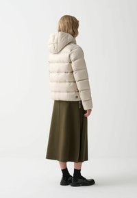 Vadderad beige jacka med huva, med en fyrkantig design och horisontell sömnad. Kombinerad med en lång grön kjol och svarta skor.