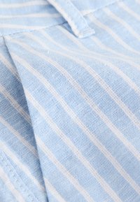 Svetlomodro blago z belimi navpičnimi črtami; vidna je teksturirana tkanina, ki daje mehak in zračen videz.