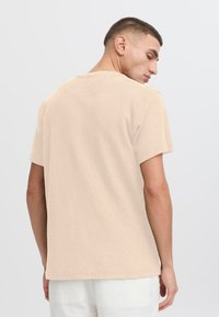 Beige T-Shirt mit kurzen Ärmeln und strukturiertem Stoff, mit einem Rundhalsausschnitt und einer lockeren Passform, von hinten betrachtet.