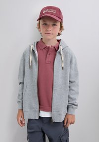 Šedá mikina na zip s krémovými šňůrkami přes vínový polo tričko. Vínová baseballová čepice s bílým logem. Neformální, volný styl.