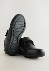 Chaussures en cuir noir avec une sangle en velcro et une semelle en caoutchouc texturée arborant des rainures horizontales. La marque "next" est visible sur la semelle.